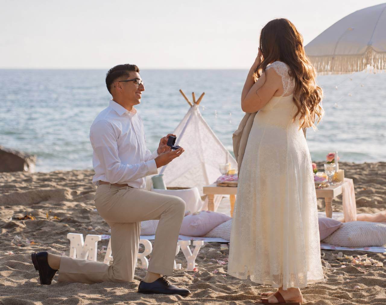guide-for-proposals-golden-hour-beach-proposal Golden hour beach proposal with ocean backdrop, cream dress, neutral trousers, picnic styling, and ring box for a guide for proposals