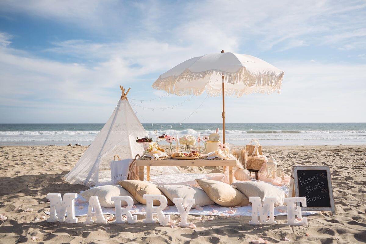 Luxury beach proposal picnic with MARRY ME letters, umbrella, flowers, and rose petals
